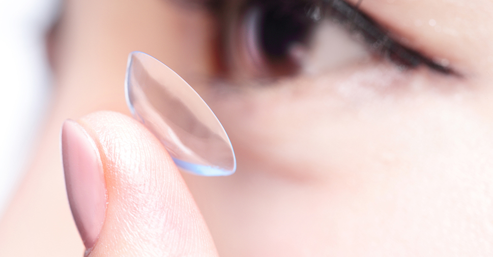 A closeup of a lady holding a contact lens on her index finger. A side profile view of her face is in the background.
