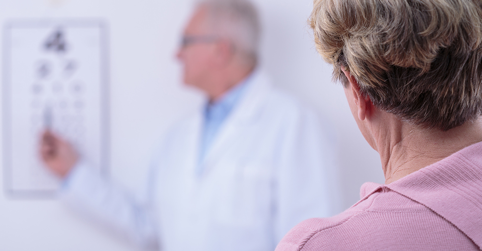 Showing back of a lady's head and shoulders staring at a vision test with a doctor pointing towards letters/numbers.