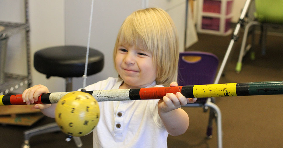 A smiling young boy during a vision therapy session, holidng a bat up near a ball that is hanging from the ceiling.